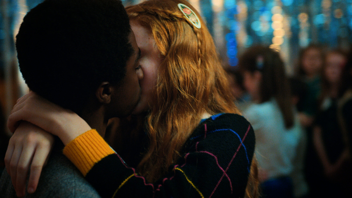 Two teenagers share a kiss in a close embrace at a school dance, surrounded by other students and festive blue decorations in the background.