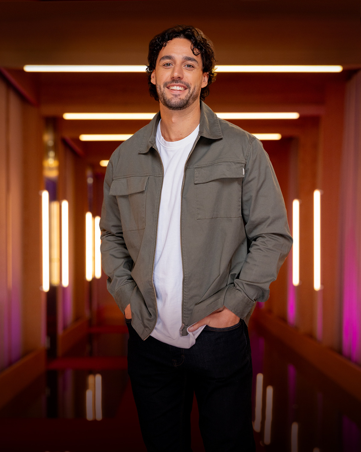 Man with curly hair wearing a green jacket and white t-shirt stands smiling in a modern hallway with bright vertical lights and pink-purple ambient lighting in the background.