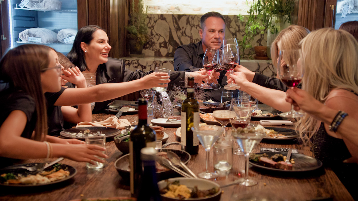 Group of people enjoying a celebratory dinner at a restaurant, raising their glasses for a toast around a wooden table set with plates of food, drinks, and wine bottles in an elegant, warmly lit indoor setting.