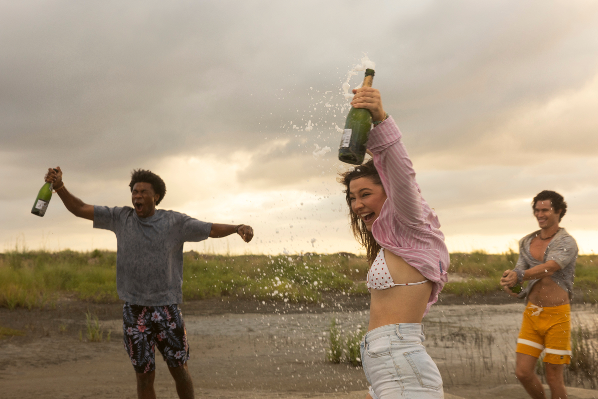 Three young adults outdoors on a beach or marsh, celebrating and spraying champagne, wearing swimsuits and casual clothes, with cloudy skies and grassy surroundings in the background.