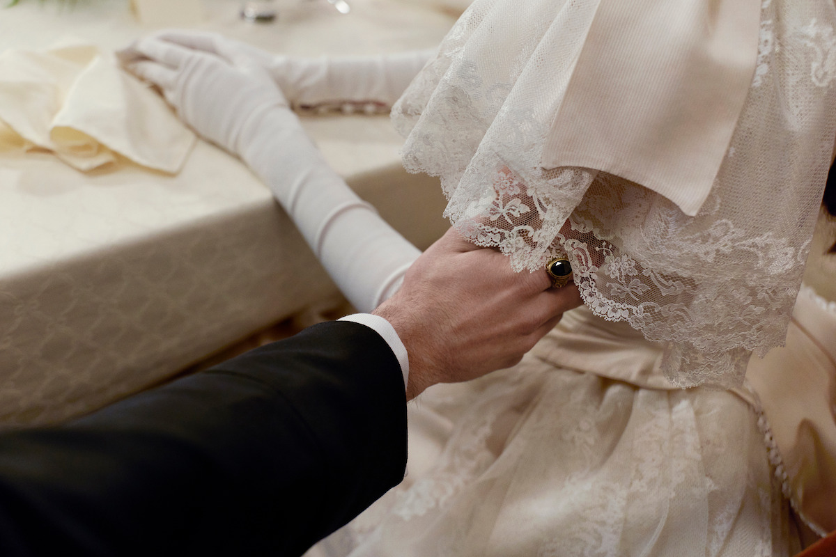 A person in formal attire gently holds the arm of someone in an elegant lace wedding dress and gloves at a table, evoking a tender, romantic atmosphere at a wedding or special event.