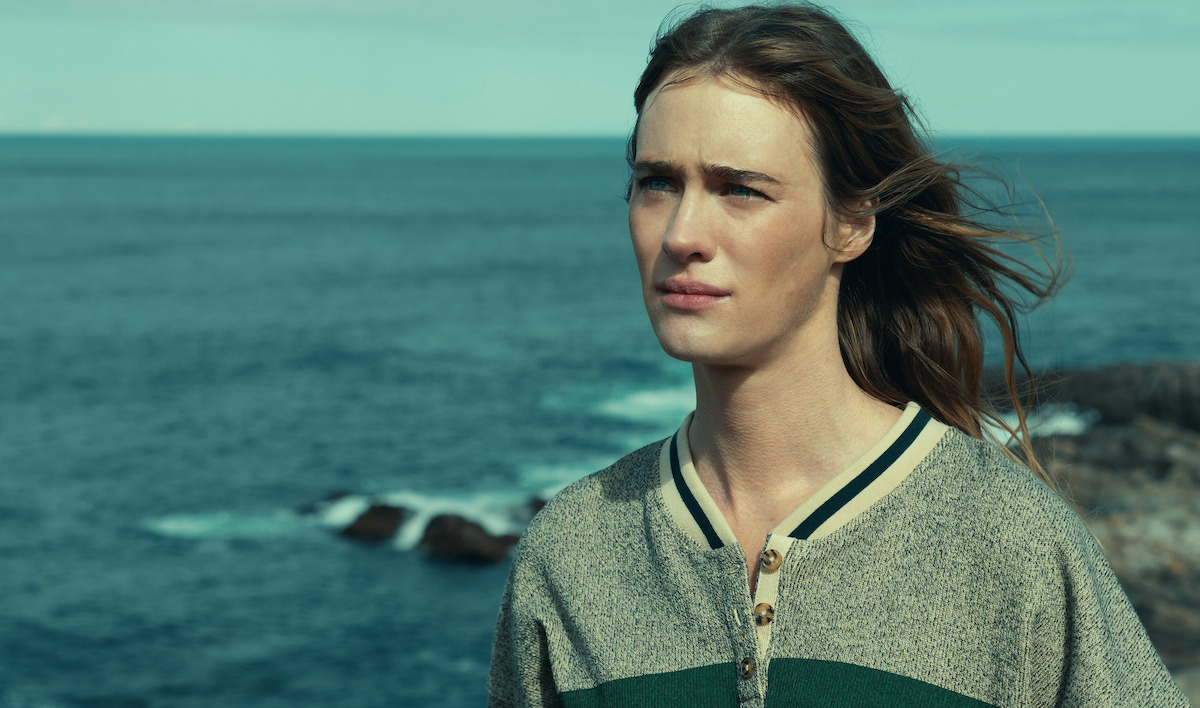 Person with long hair standing outdoors near the ocean, wearing a gray and green sweater. The sea and rocky shore are in the background under a clear sky, creating a serene coastal scene.