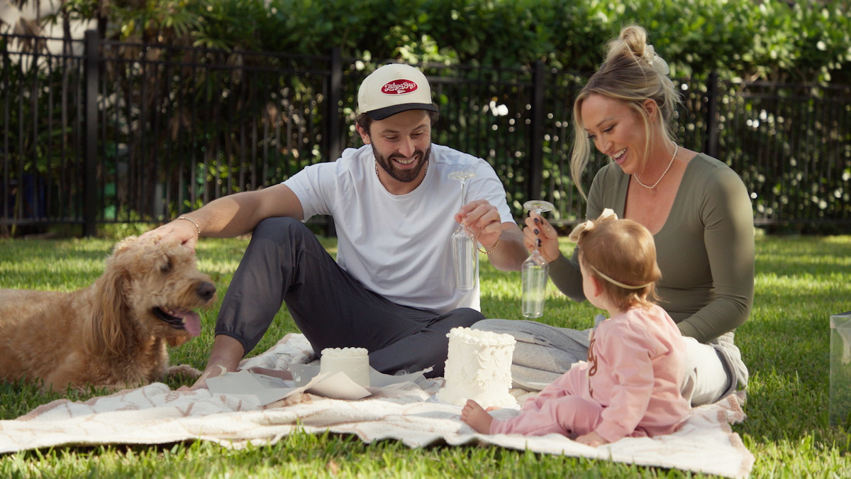 A man, woman, and toddler have a picnic on a blanket in a sunny backyard with a happy dog nearby, surrounded by grass, trees, and a black iron fence, enjoying time together with snacks and drinks.