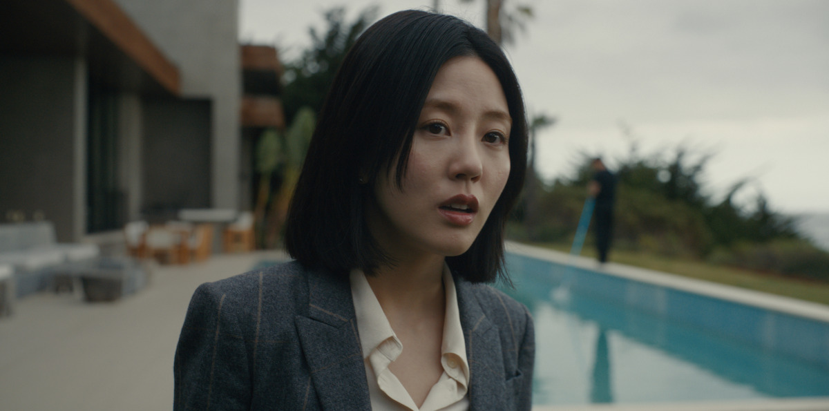 A woman in a suit stands outside by a modern house and pool, looking concerned. The setting is outdoors with patio furniture, greenery, and a person in the background near the pool under a cloudy sky.