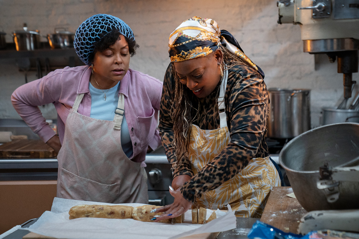 Two women in aprons prepare dough in a busy kitchen, one slicing while the other observes; shelves with pots and an industrial mixer are visible, creating an active, collaborative baking environment.