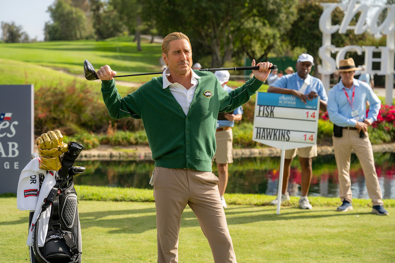 Golfer in green cardigan poses with club on shoulders at golf course, with caddie and officials nearby, score sign and lush landscape in background, sunny outdoor tournament scene.