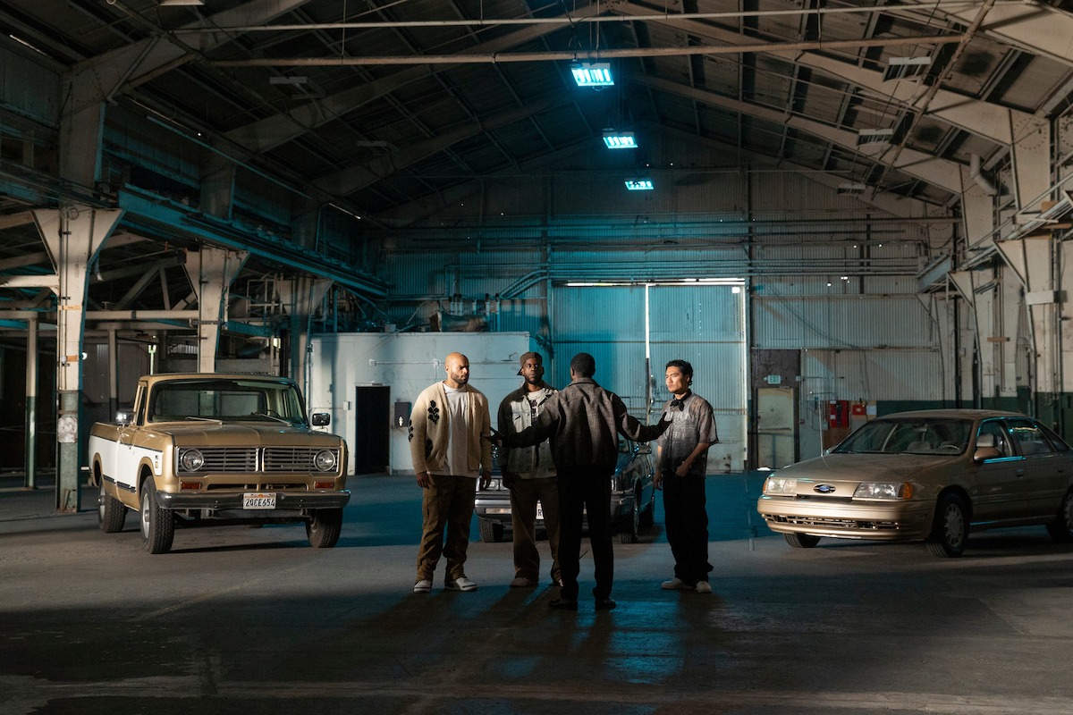 Five men standing and talking inside a large, industrial warehouse with high ceilings, flanked by two parked cars—a vintage SUV and a sedan—under artificial lighting.