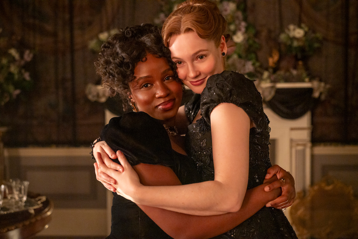 Two women in elegant period dresses embrace and smile warmly in a lavishly decorated room with floral arrangements and vintage decor, creating a joyful and affectionate atmosphere.