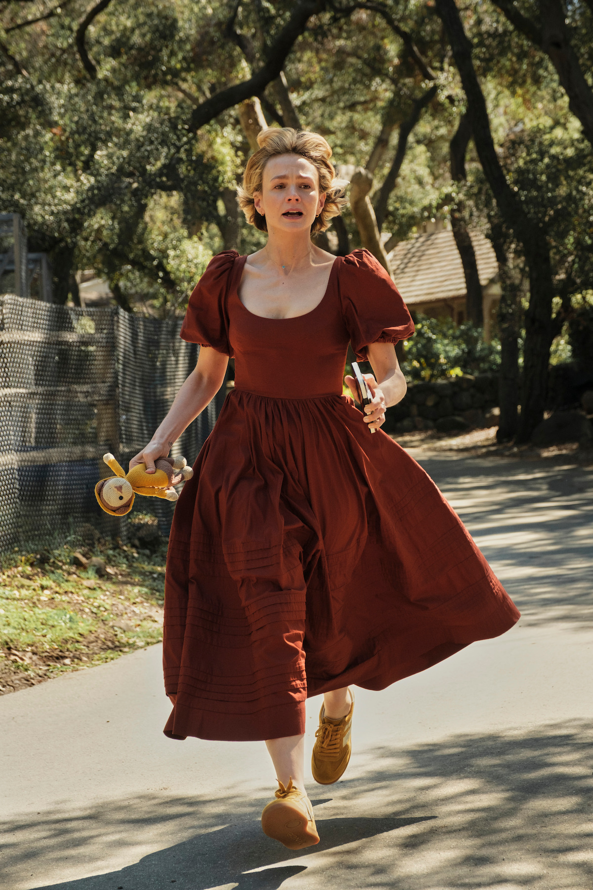Woman in a flowing red dress runs on a sunlit outdoor path, holding children’s toys, with trees and a fence in the background, suggesting urgency or concern in a rural or suburban environment.