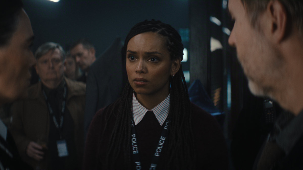 A woman with braids wearing a police lanyard stands indoors, looking concerned, surrounded by colleagues in a dimly lit, busy police station environment.