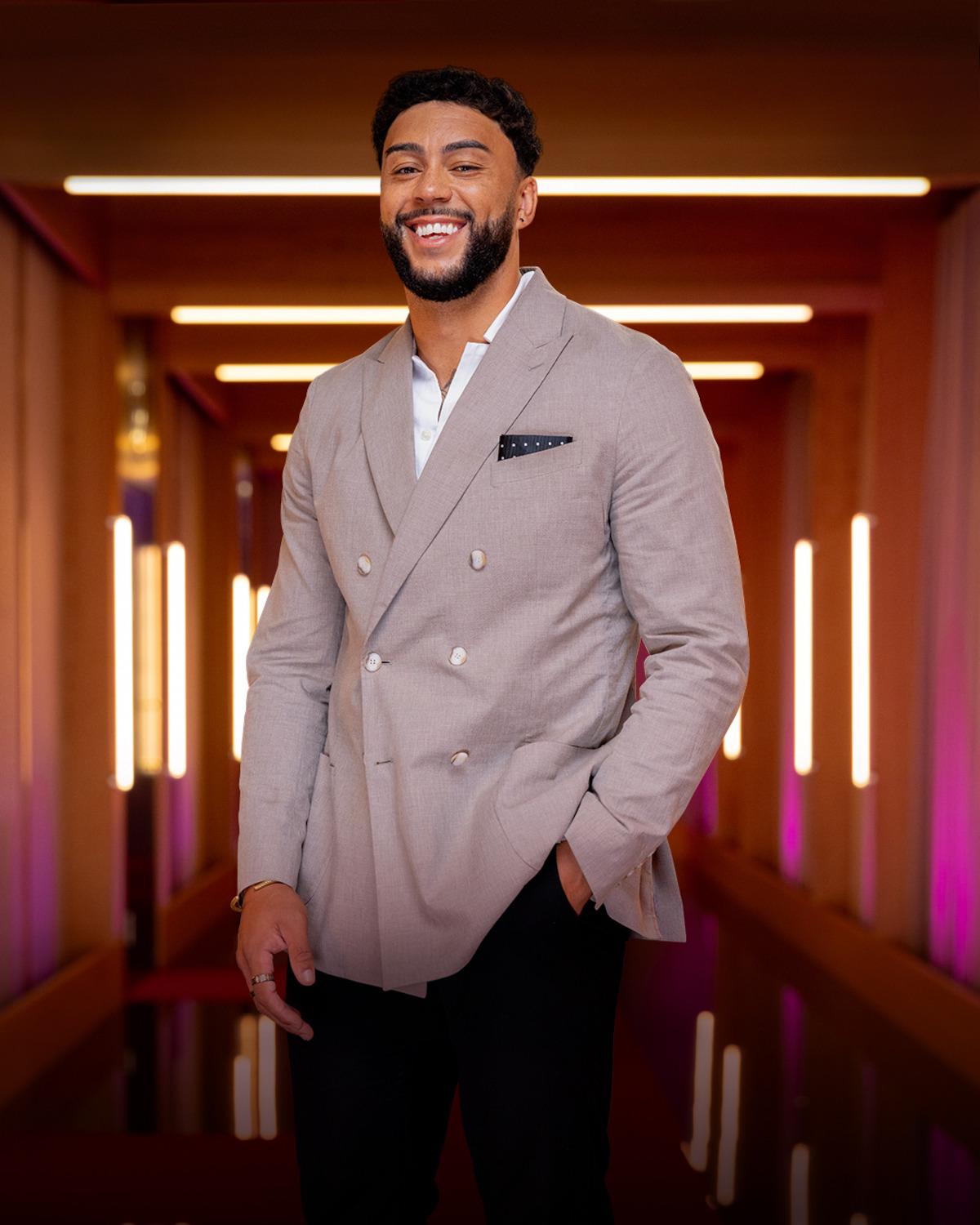 Man in a light beige suit jacket and black pants standing in a stylish, warmly lit hallway with glowing vertical lights and a modern interior design, smiling with one hand in his pocket.