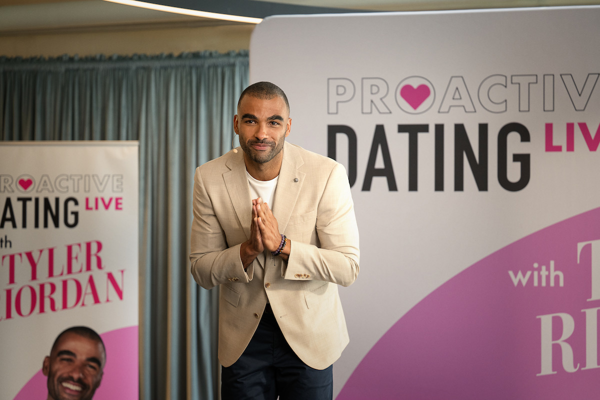 Man in a beige blazer stands with hands together on stage at a "Proactive Dating Live" event, banners and curtains in the background, event setting appears professional and welcoming.