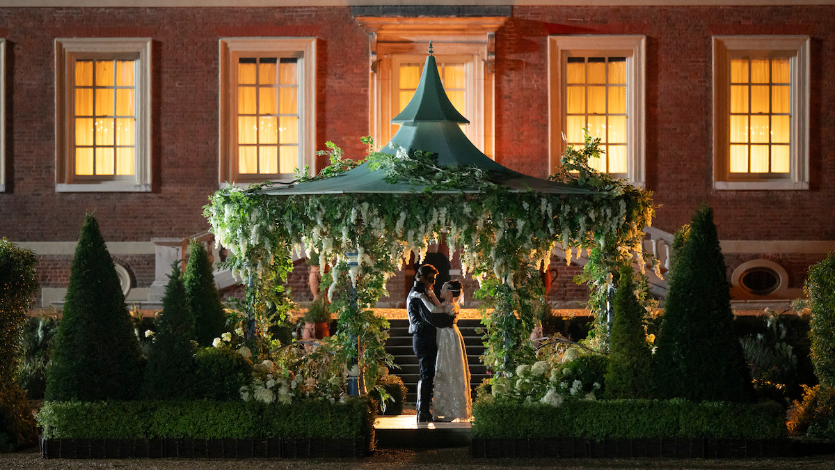 A couple embraces under a flower-decorated gazebo in a formal garden at night, with a large brick building and warmly lit windows in the background, surrounded by trimmed hedges and greenery.