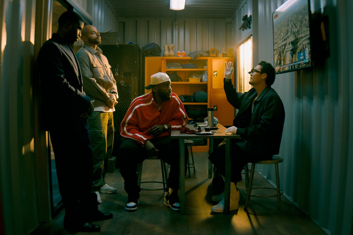 Four men in a small, dimly lit room, three standing and one sitting at a table raising his hand; shelves and lockers in the background indicate a storage or meeting space.
