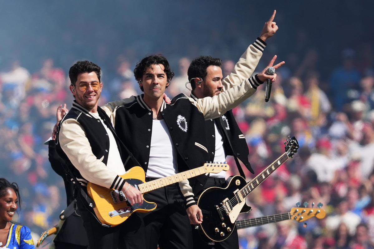 Three musicians in matching jackets perform on an outdoor stage with electric guitars, singing to a large crowd in a stadium filled with colorful spectators.