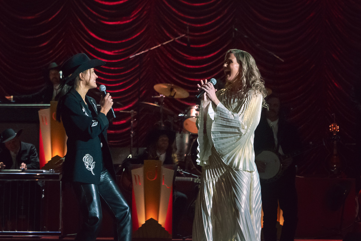 Two singers performing on stage, one in a black outfit with a hat and the other in a white dress, with a live band playing instruments in the background and dramatic stage lighting.