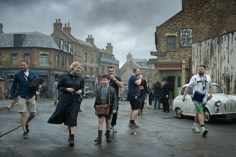 People walk on a wet street in a historic town setting, with vintage buildings, an old car, and overcast skies. A woman and a boy in period clothing are surrounded by crew members, suggesting a film production environment.