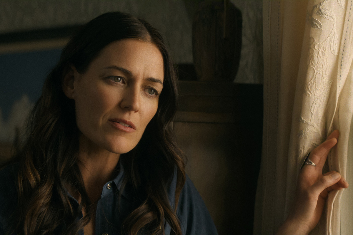 Woman with long brown hair in a blue shirt stands indoors near a window with lace curtains, looking out thoughtfully while gently holding the curtain. Warm, soft lighting creates a calm and intimate atmosphere.