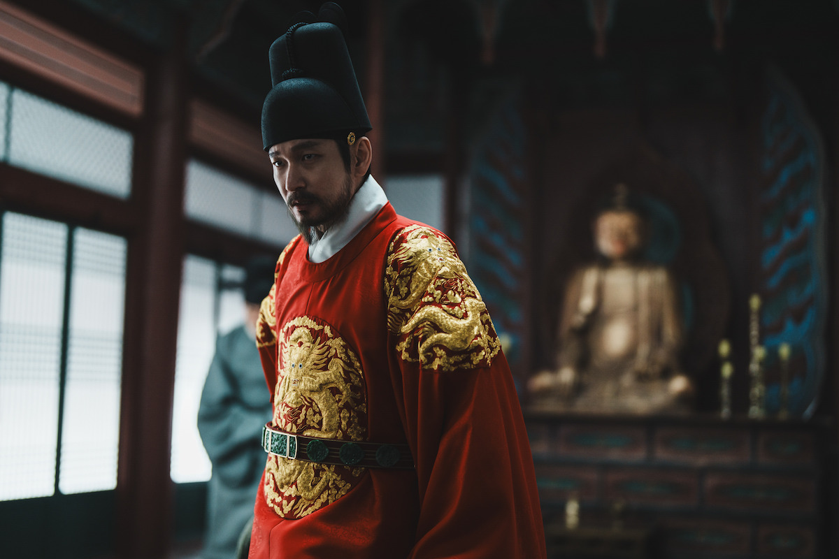 Man dressed in traditional royal Asian attire stands in an ornate room with wooden details and a large golden Buddha statue in the background, suggesting a historical or ceremonial setting.Man dressed in traditional royal Asian attire stands in an ornate room with wooden details and a large golden Buddha statue in the background, suggesting a historical or ceremonial setting.