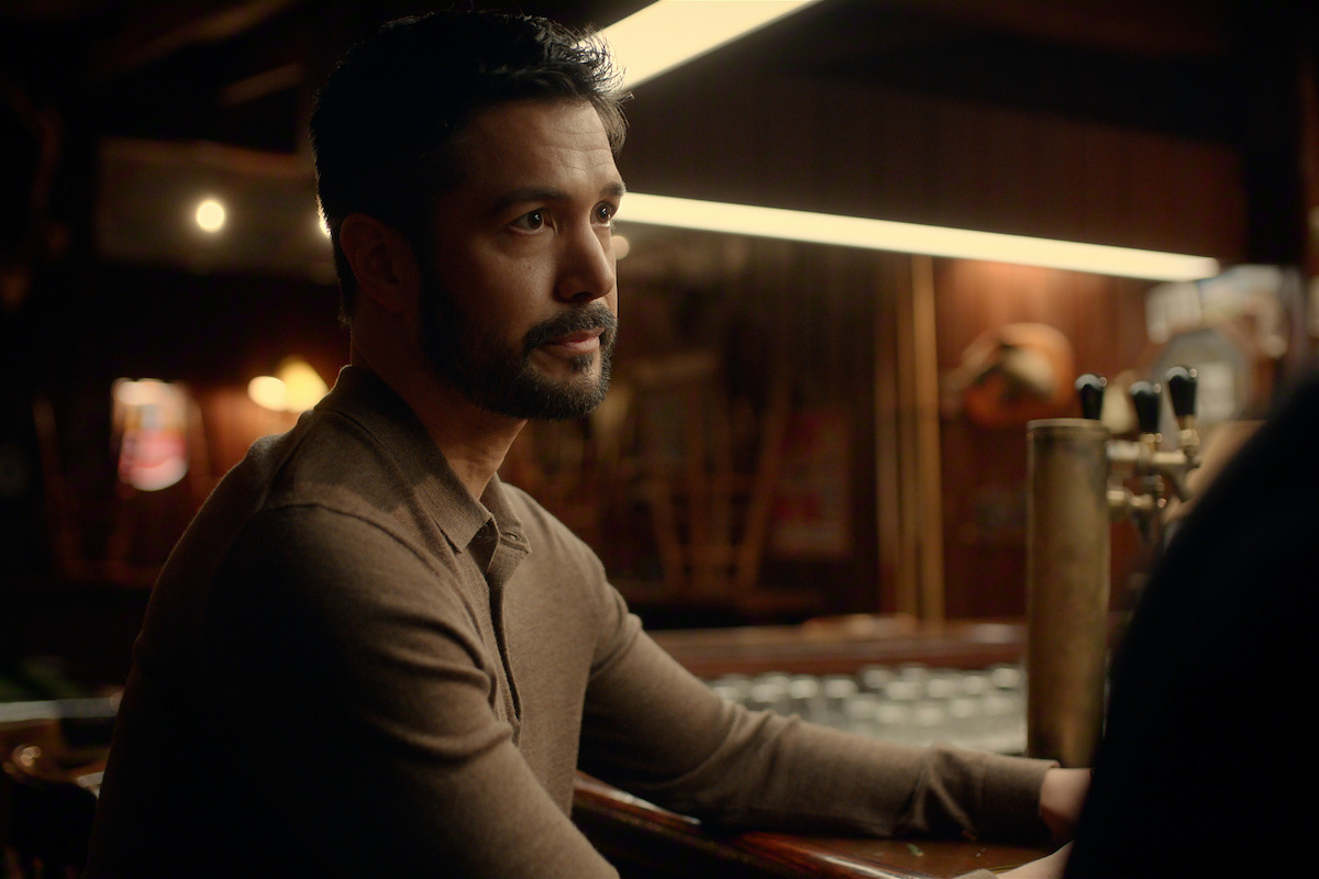 Man with a beard sitting at a dimly lit bar, wearing a brown shirt, looking thoughtful; background features warm lighting, wooden walls, and bar equipment.