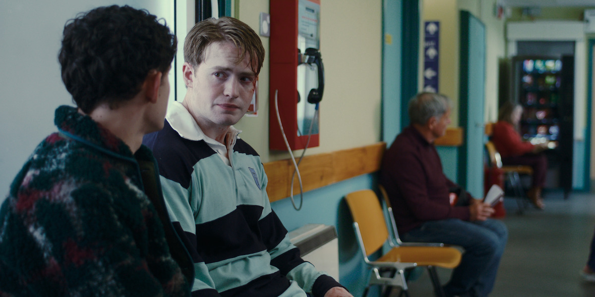 Two young men sit and talk in a hospital hallway, with other people waiting on yellow chairs; a payphone and vending machine are visible in the background, creating a somber atmosphere in a medical environment.