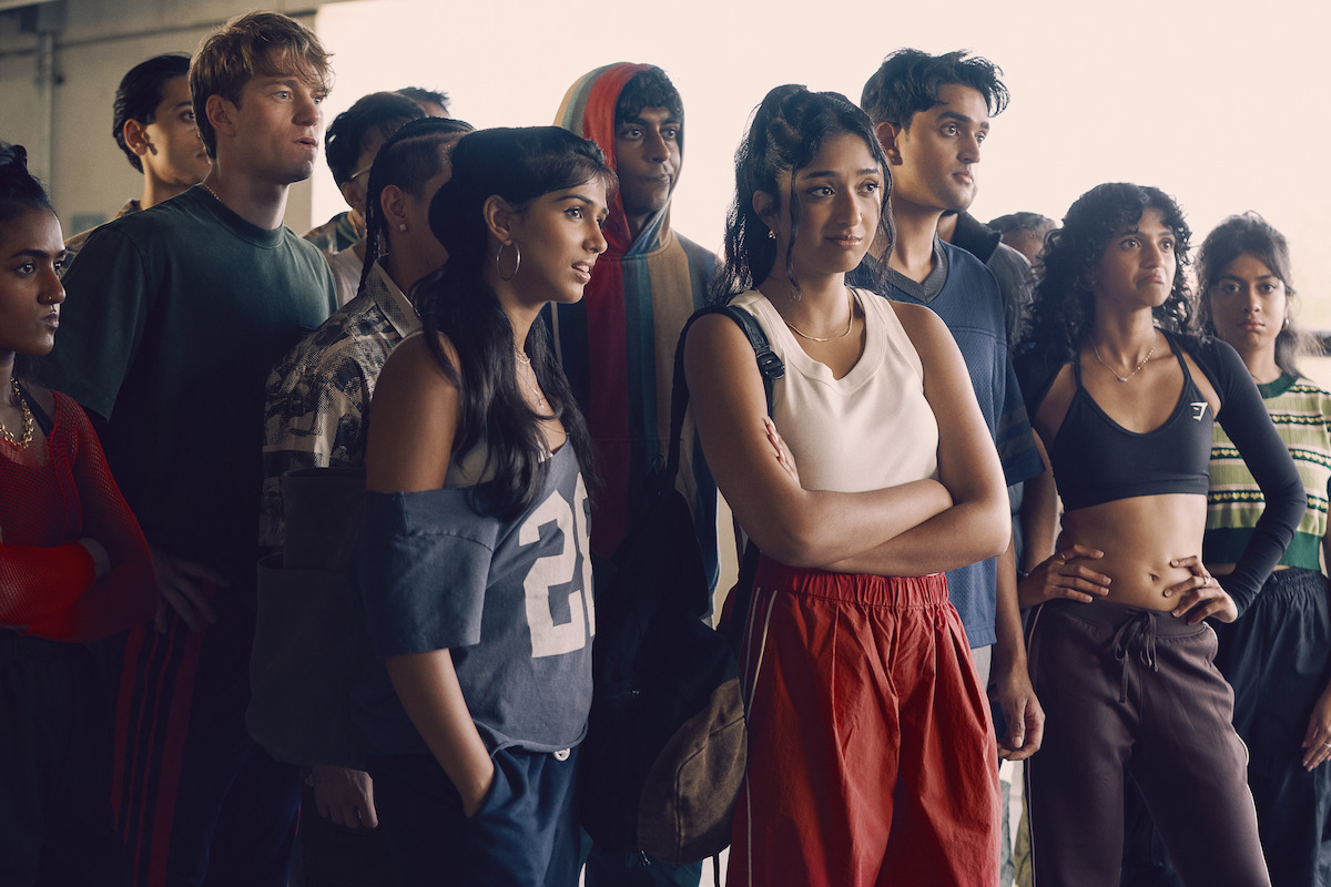 A diverse group of young adults in sporty, casual clothing stand together indoors, appearing focused and attentive, possibly listening to someone or watching an event in a well-lit urban setting.