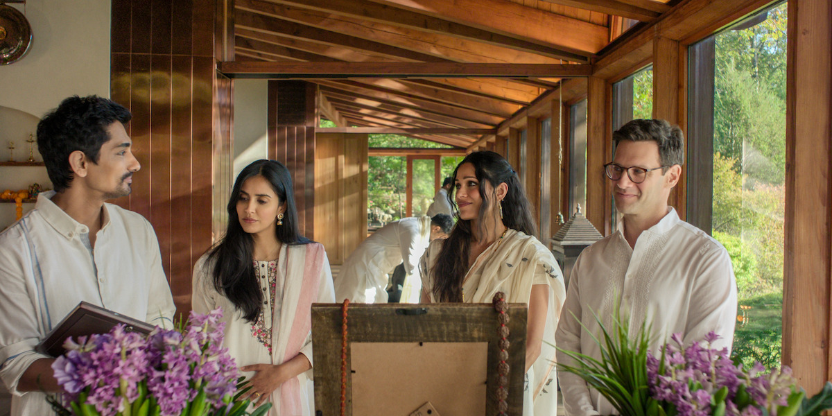 Four people in light-colored clothing stand indoors by large windows with greenery outside, surrounded by purple flowers, engaging in conversation in a warm, sunlit setting with wooden beams and soft natural light.
