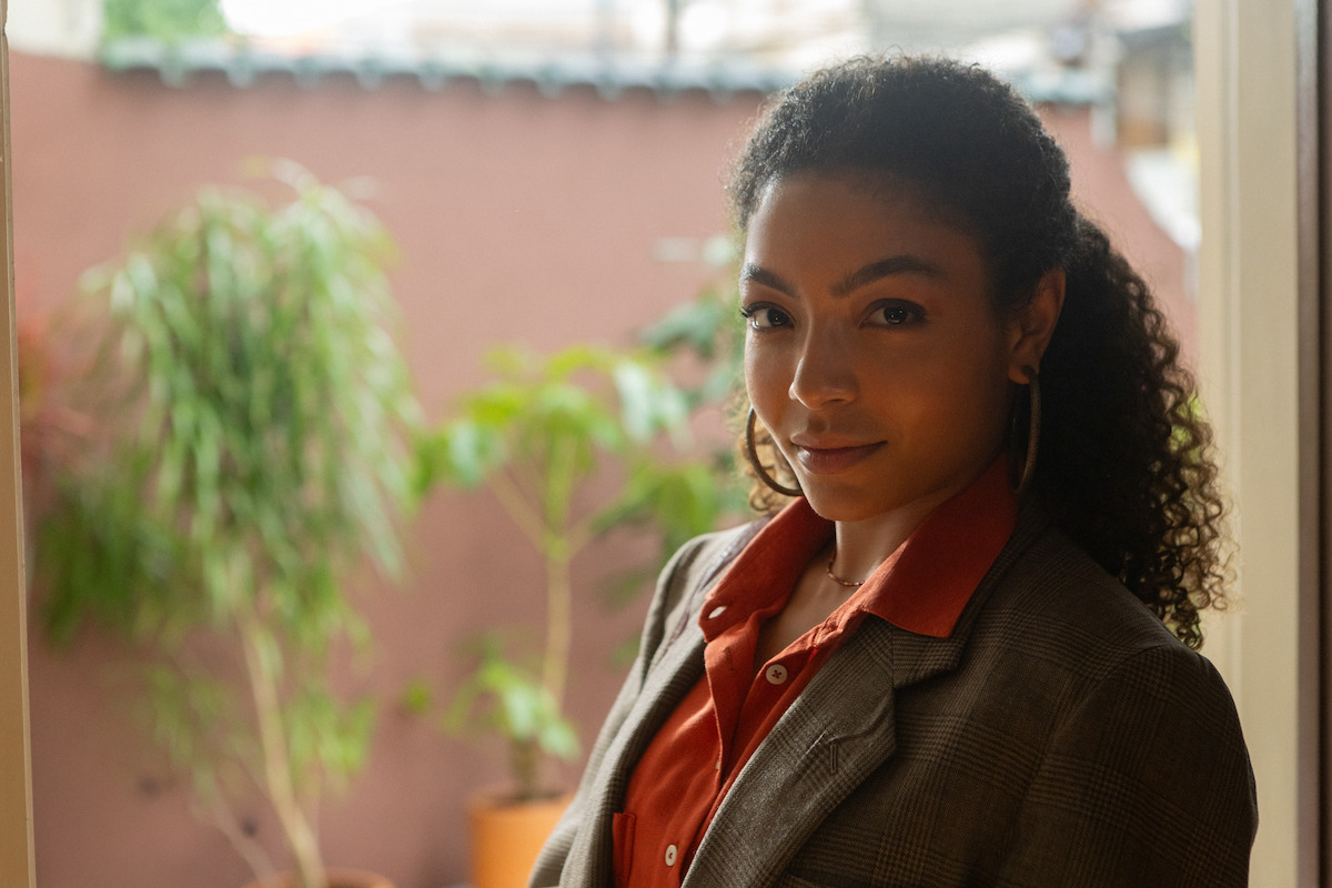 Mulher negra está de terno e camisa laranja com plantas no plano de fundo