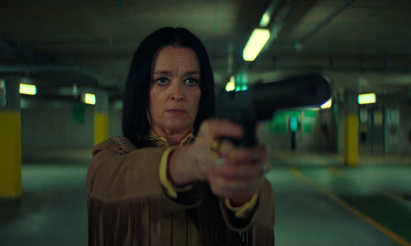 A serious woman in a fringe jacket aims a silenced pistol in an underground parking garage, creating a tense and suspenseful atmosphere.