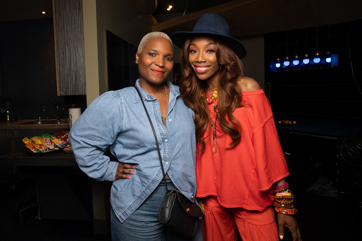 Two women posing and smiling indoors, one in a denim outfit with a crossbody purse, the other in a red outfit and blue hat, with snacks and dim lighting in the background.