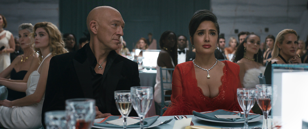 Elegant man and woman seated at a formal event, surrounded by well-dressed guests at round tables with drinks and table settings; the atmosphere is upscale and attentive.