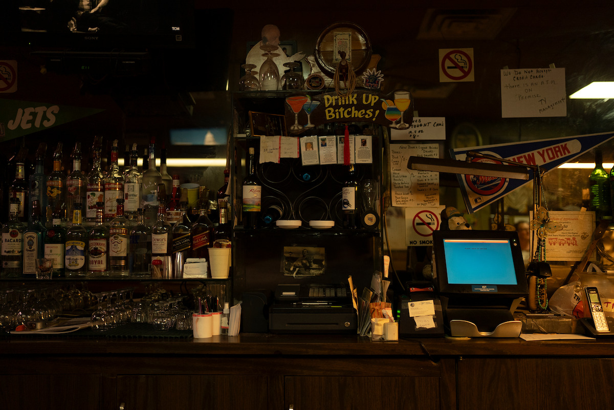 Dimly lit bar counter with bottles of liquor, glasses, a cash register, drink-making tools, handwritten notes, sports pennants, and humorous signs in a cozy, casual setting.