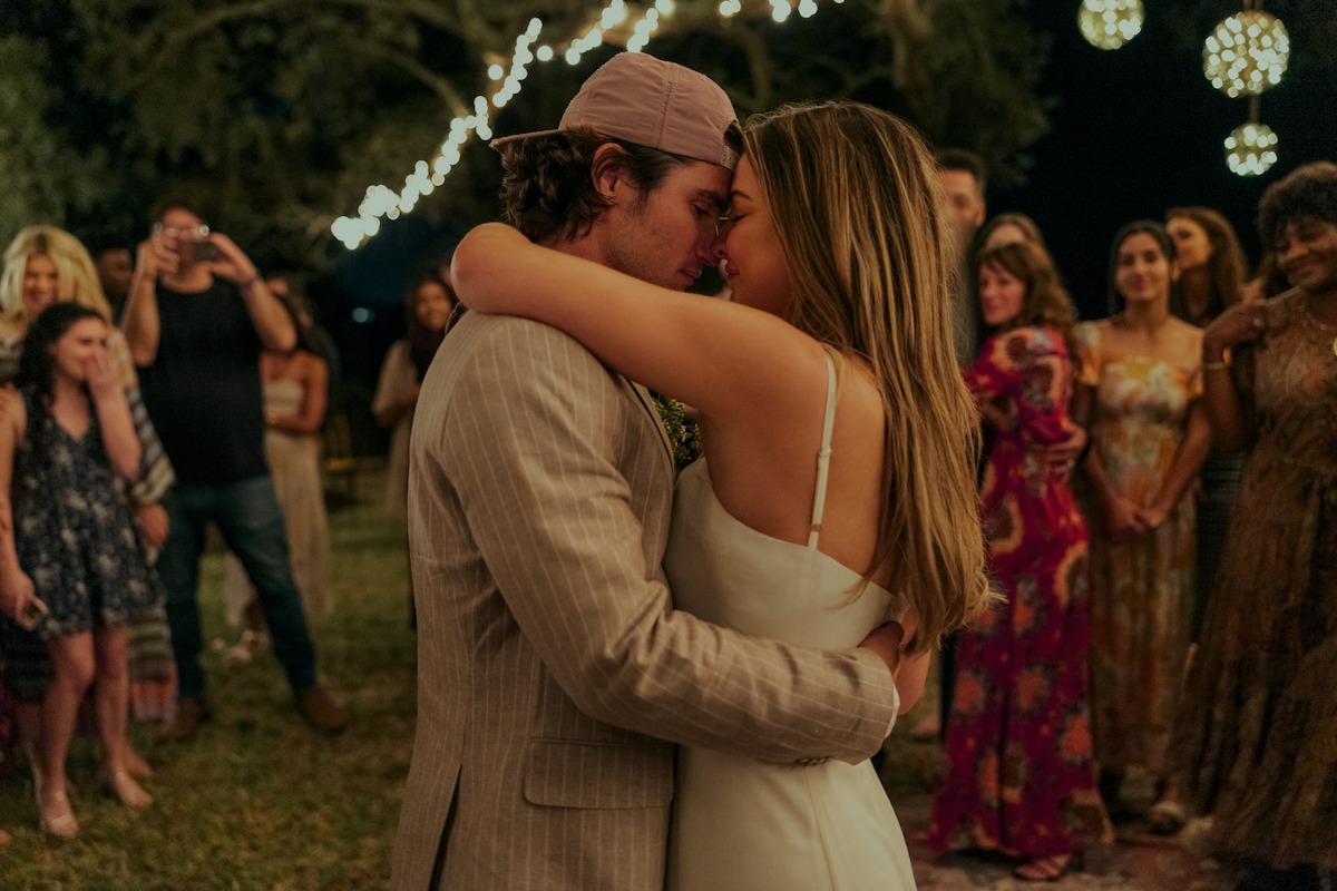 A couple shares a close embrace during a nighttime outdoor celebration, surrounded by friends and family, with string lights and hanging lanterns creating a warm, festive atmosphere.