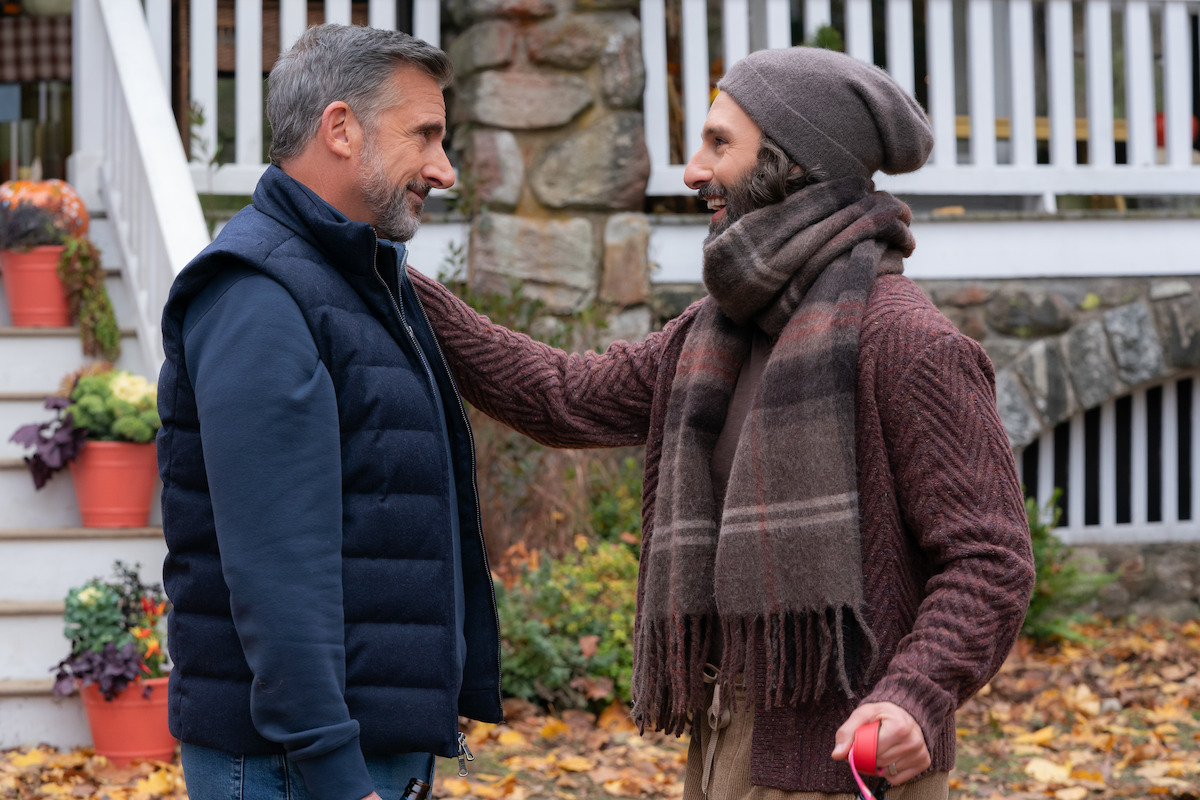 Two men warmly greet each other outdoors near a house with stone steps and potted flowers in autumn, surrounded by fallen leaves. Both are dressed in warm clothes, one wearing a scarf and hat, the other a vest.
