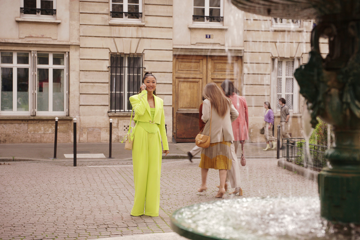 Ashley Park as Mindy stands at the Place de l’Estrapade wearing a bright yellow outfit in Season 4 of ‘Emily in Paris’