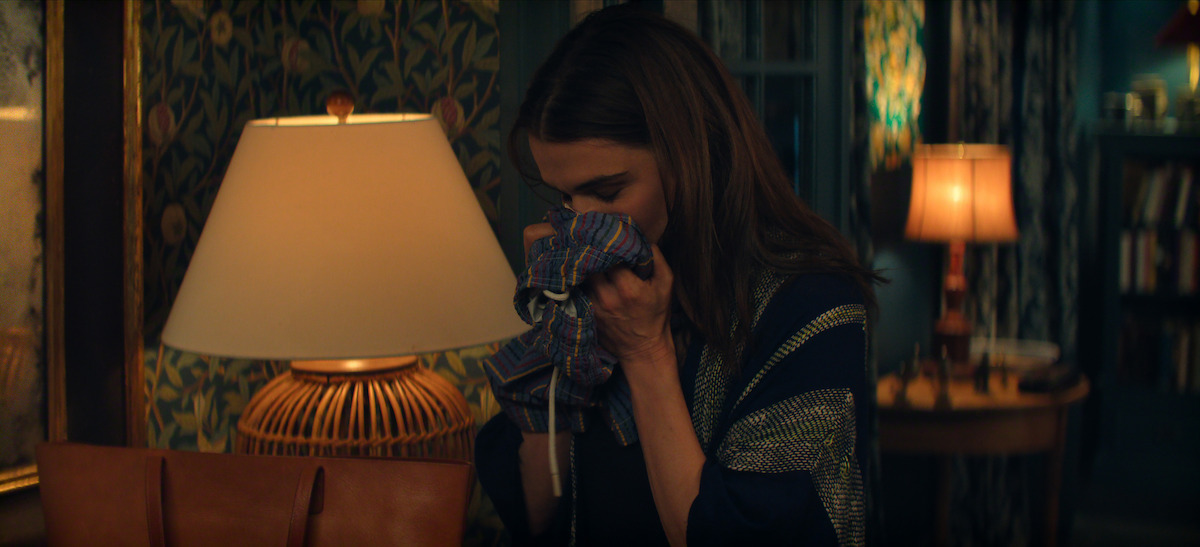 A woman stands in a warmly lit, cozy room with patterned wallpaper and bookshelves, holding and smelling a piece of clothing, with two table lamps illuminating the space.