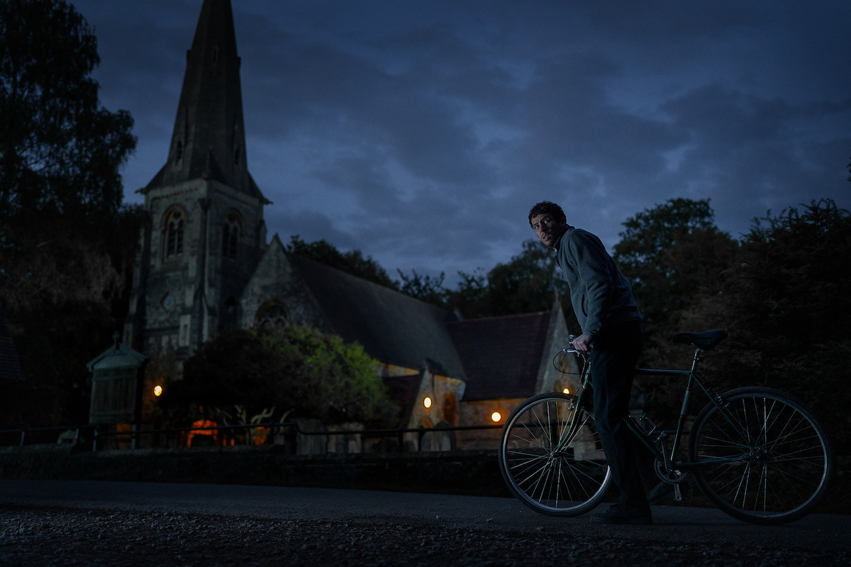 A man with a bicycle stands on a road at dusk in front of a lit, old stone church with a tall steeple, surrounded by trees. The moody, dark blue lighting creates a tense, mysterious atmosphere.