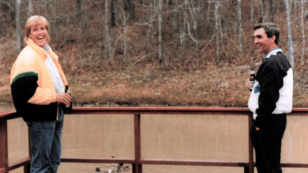 John Elway and Mike Shanahan stand on a wooden deck over a pond, holding drinks and smiling, with leafless trees and a forest in the background on an overcast day.