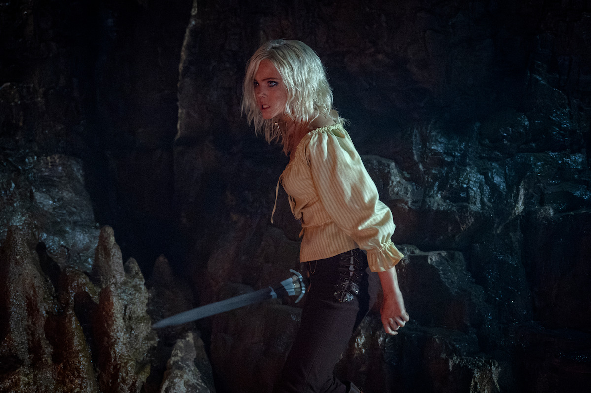 A woman with blonde hair holds a sword, standing in a dark, rocky cave. She looks tense and alert, wearing a light blouse and dark pants. The environment is moody and suspenseful, evoking a sense of danger and adventure.