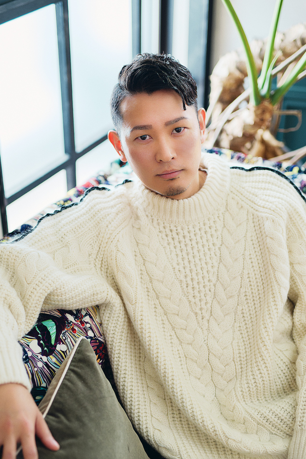 Man in a cozy white knit sweater sits on a patterned couch by a sunny window, surrounded by plants, creating a relaxed and comfortable indoor atmosphere.
