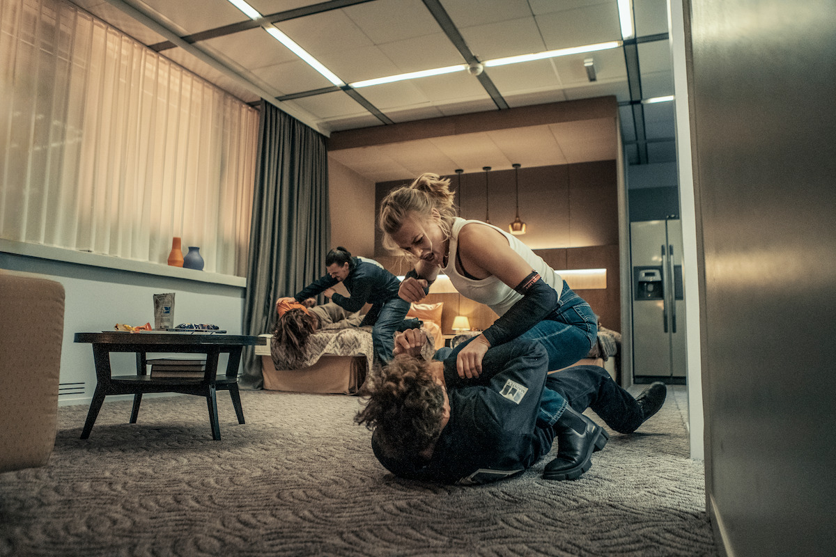 Jeanne Goursaud as Sara Wulf holds down a man on a carpeted apartment room floor in an image from the film ‘Exterritorial.’