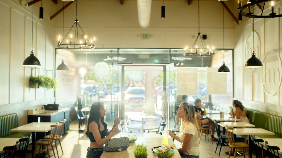 Two women sit at a table eating and talking in a bright, modern cafe with large windows, chandeliers, green plants, and other diners; sunlight pours through the entrance, creating a welcoming atmosphere.