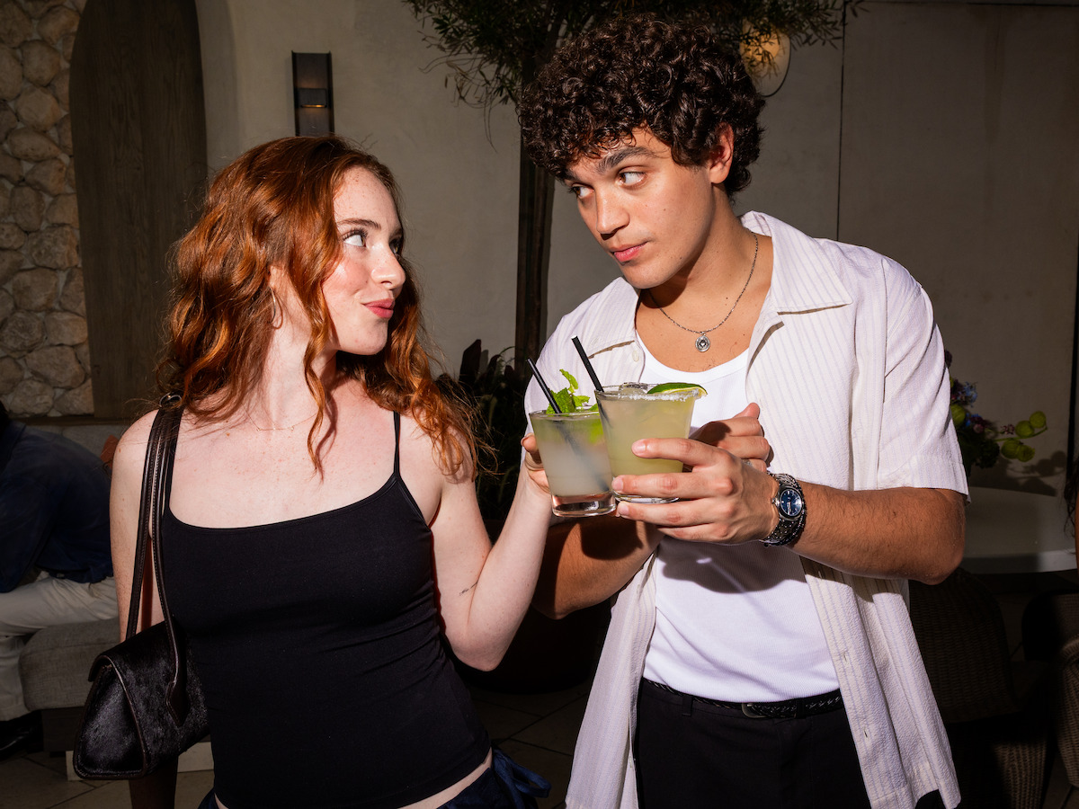 Two young adults clinking drinks, smiling at each other in a cozy indoor setting with plants and stone walls, dressed casually, suggesting a relaxed social gathering or night out.