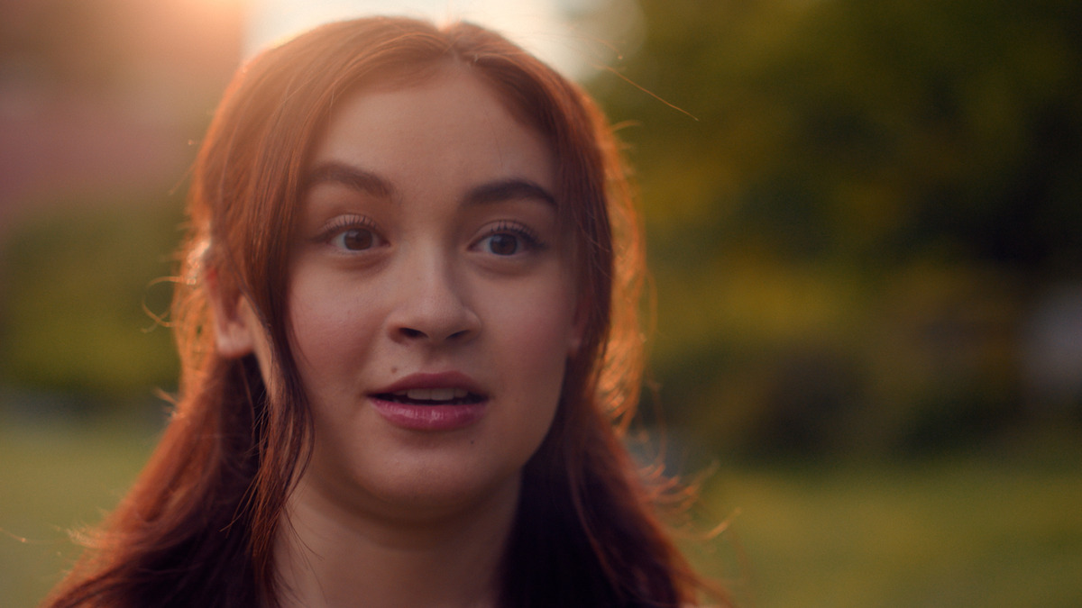 A young woman with long brown hair looks surprised outdoors in a park or garden setting during sunset, with soft natural lighting and a blurred green background.