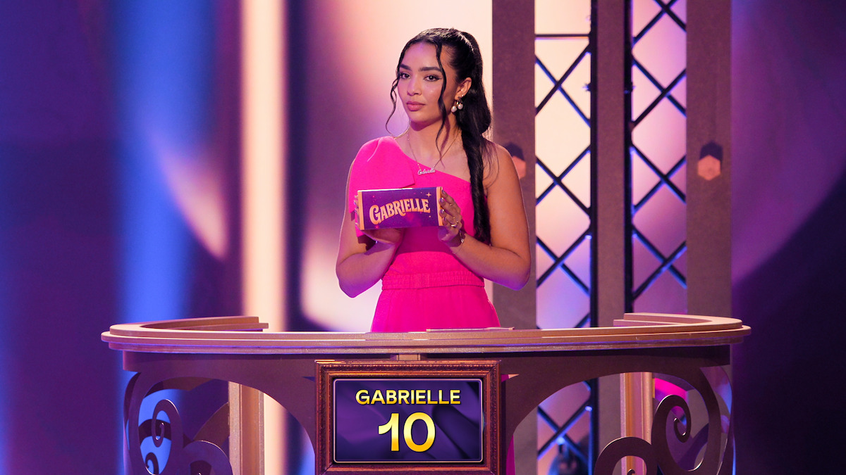 A woman in a pink dress stands at a game show podium labeled "Gabrielle 10" holding a card, with a colorful, lit studio background and decorative panels behind her.