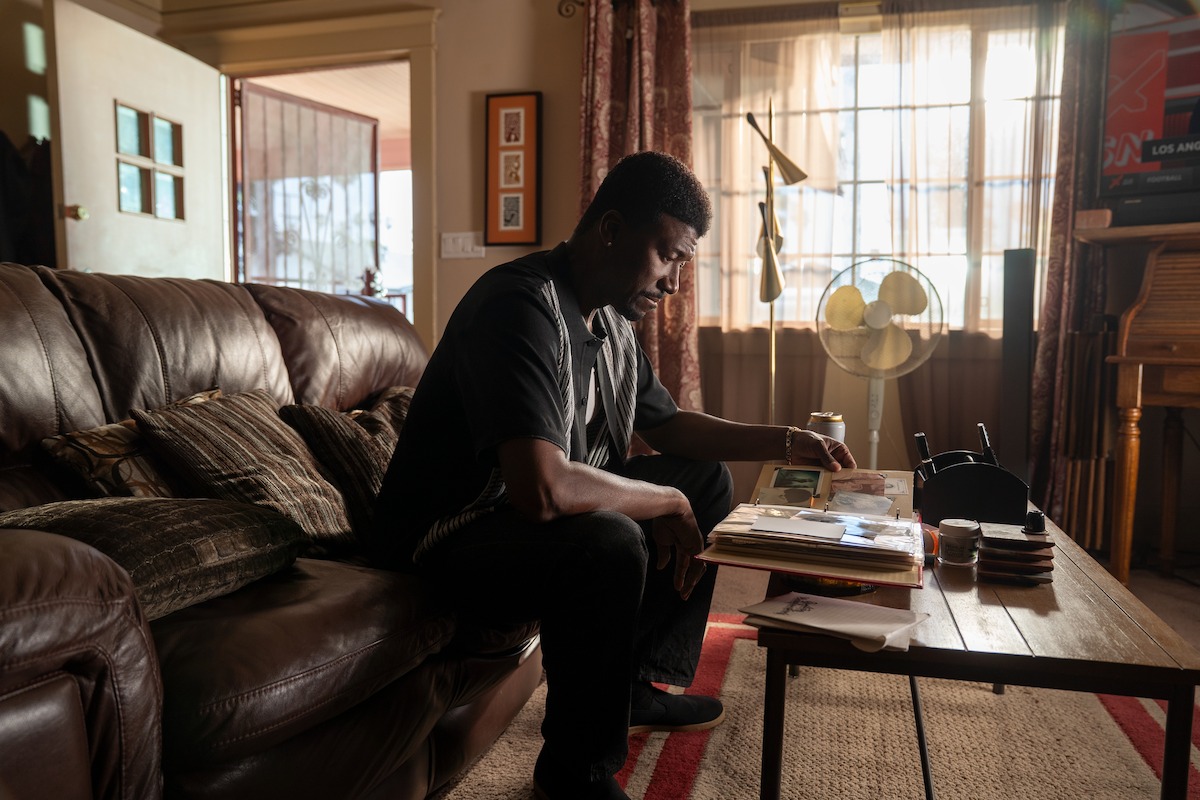 A man sits on a brown leather couch in a living room, looking at papers and photographs on a coffee table. Sunlight filters through curtains, and the room appears cozy with a fan and various household items visible.
