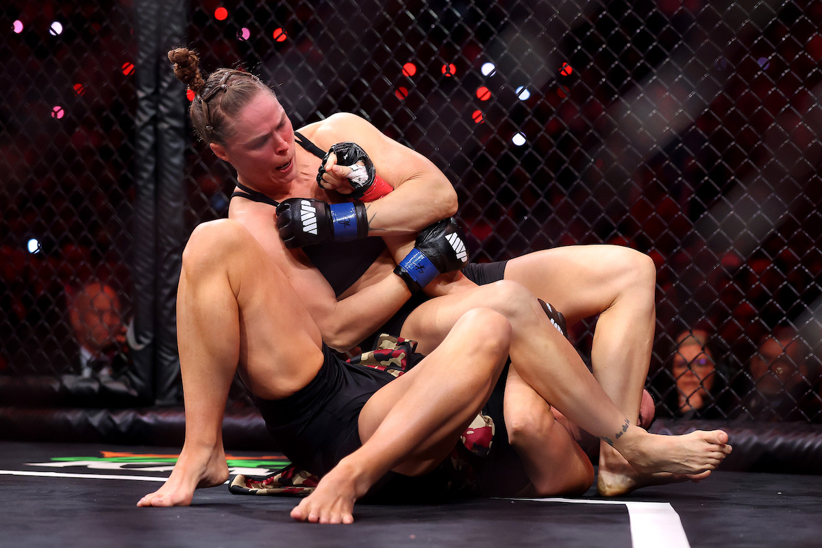 Two female MMA fighters in a cage during a match, one attempting a grappling submission hold on the other while surrounded by the crowd and under intense lighting.