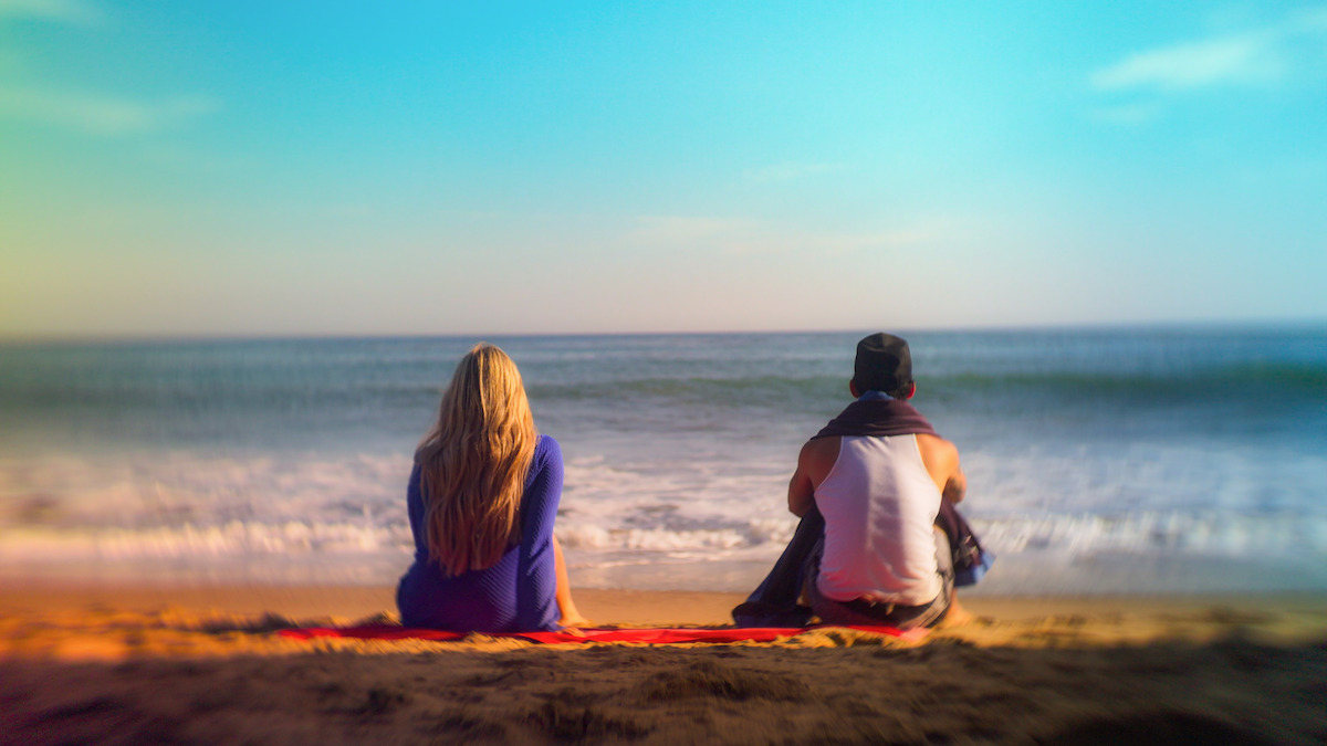 Two people sit on a blanket facing the ocean on a sandy beach under a bright blue sky, enjoying a peaceful and relaxed atmosphere.
