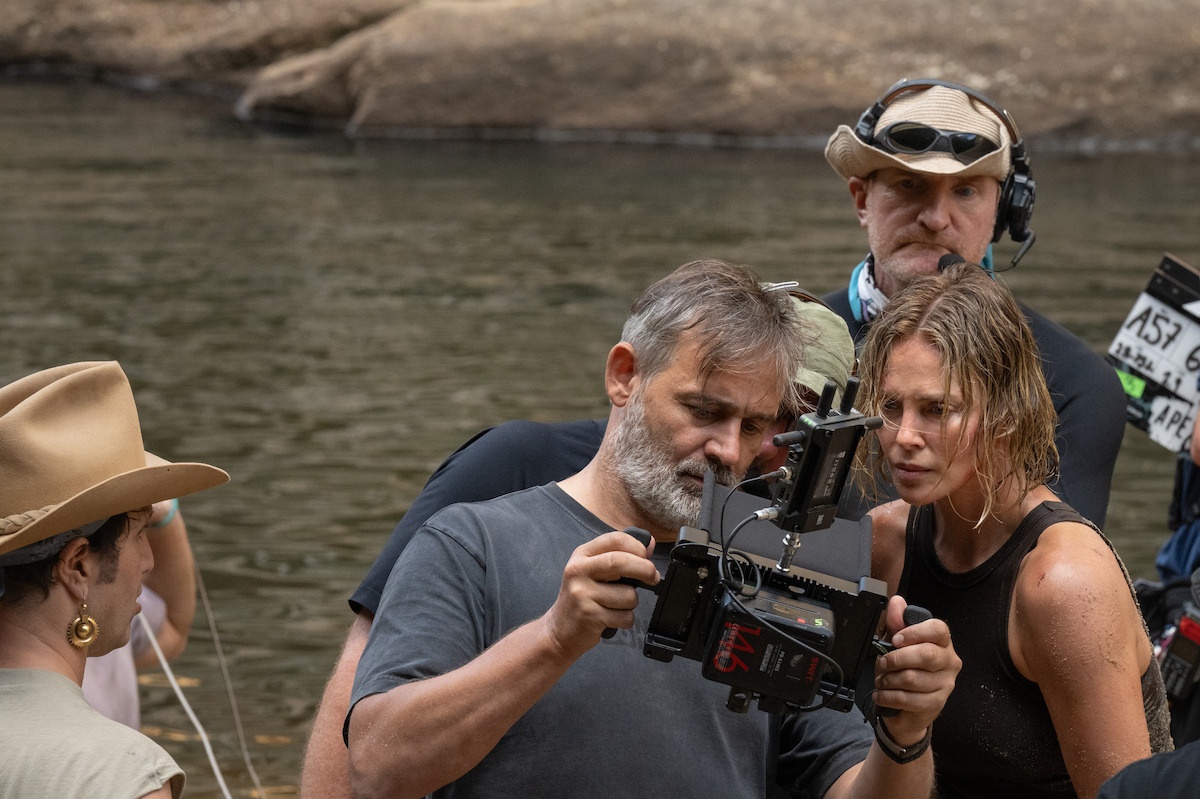 A film crew reviews footage by a river, featuring a wet actress in a black top and crew in casual outdoor gear. The mood is focused and collaborative in a natural, outdoor setting.