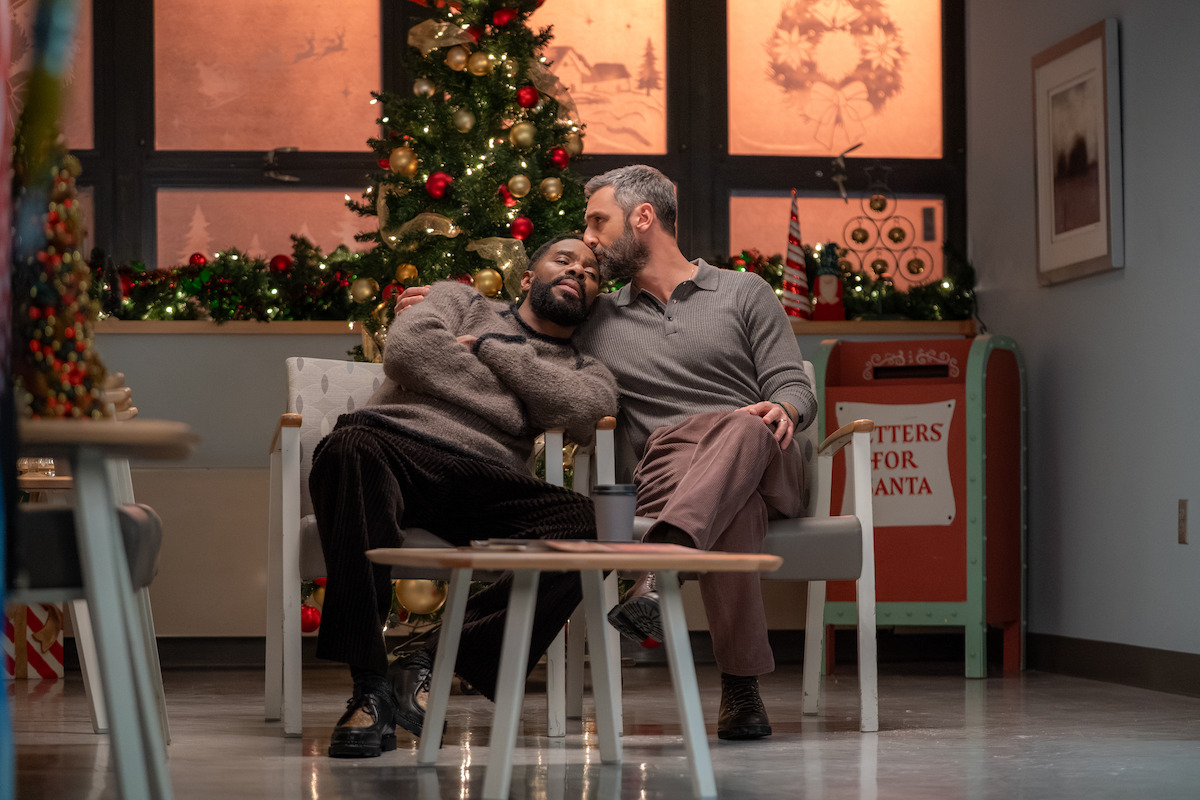 Two people sit close together in chairs in a festively decorated room, one resting their head on the other’s shoulder in front of a lit Christmas tree.